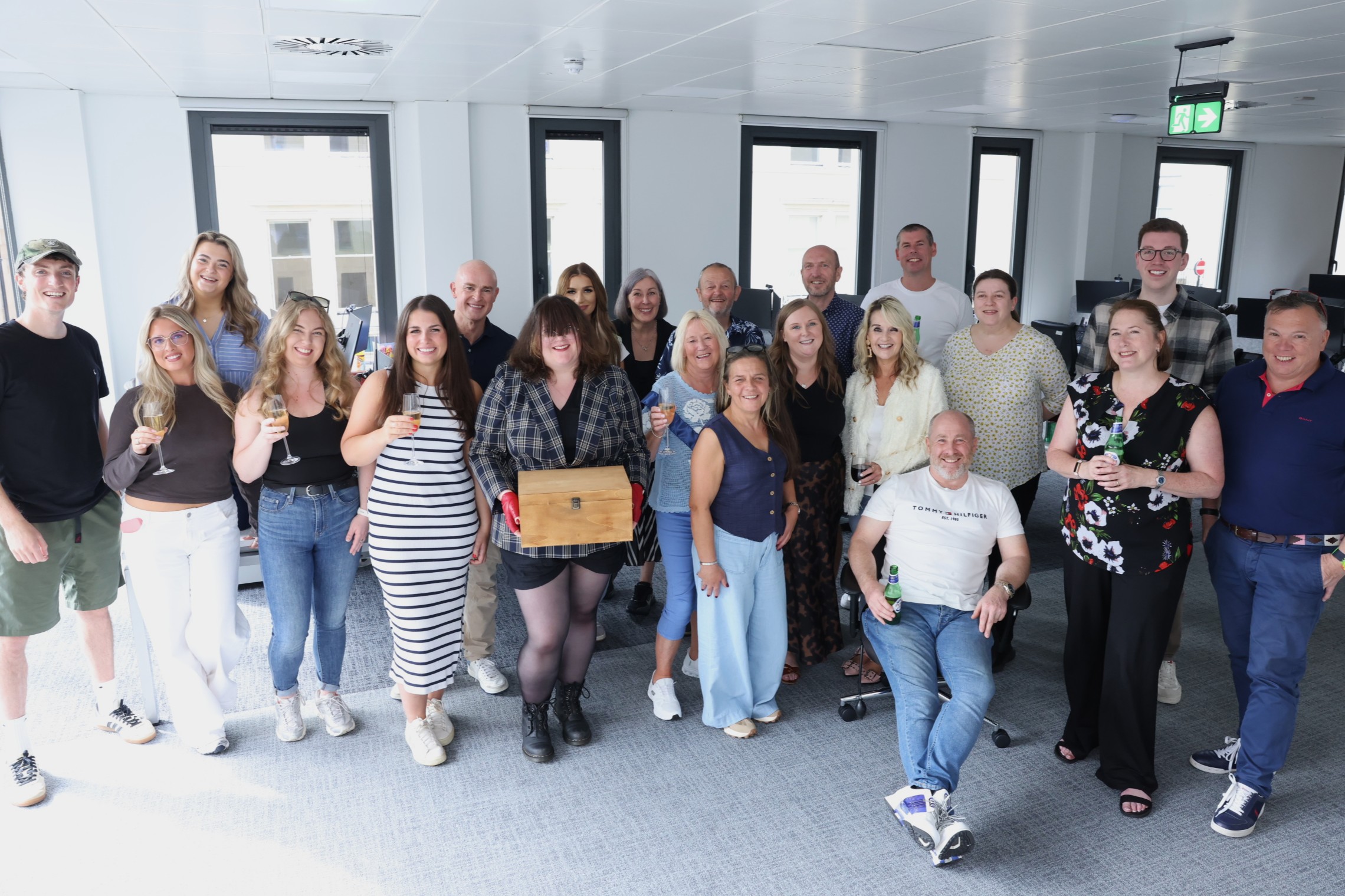 MH team gathered in a bright office for a celebration, posing together with drinks while one person stands in the centre holding a wooden presentation box