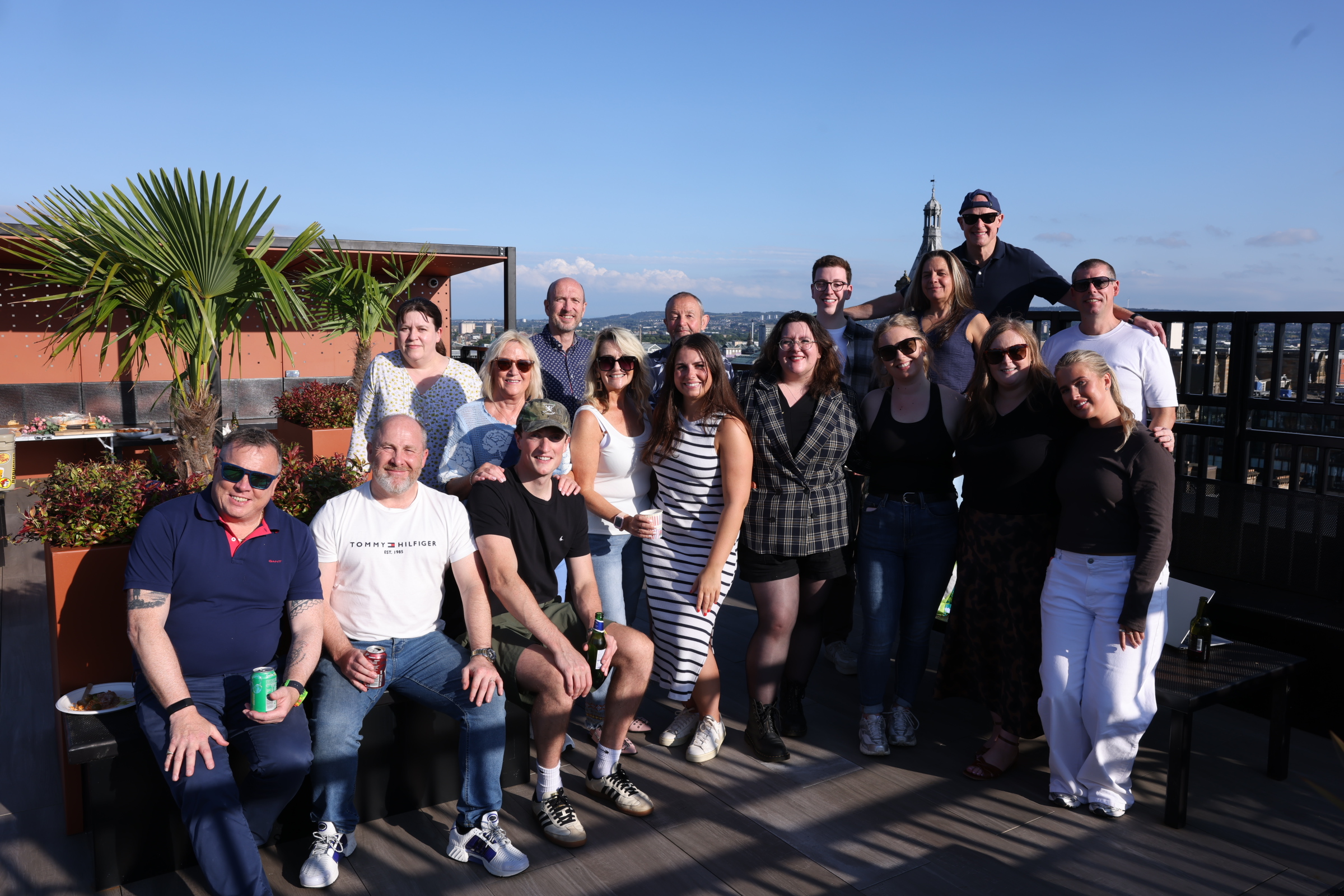 MH group photo of around 16 people gathered on a rooftop terrace on a sunny day, with potted palm trees, outdoor seating and a city skyline visible in the background.